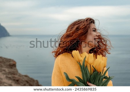 Portrait of a happy woman with hair flying in the wind against the backdrop of mountains and sea. Holding a bouquet of yellow tulips in her hands, wearing a yellow sweater