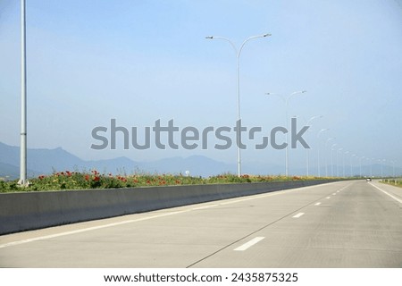 Scenic road. Straight, paved, with a concrete partition. Tall street lamps and poppies on the dividing strip. Hills and mountains on the horizon. Spirit of adventure. Forward movement. Royalty-Free Stock Photo #2435875325