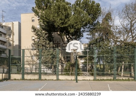 An urban basketball court with rough cement floors, a perimeter with metal fences and baskets on metal posts screwed to the ground in an area with trees and buildings
