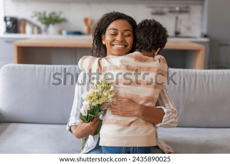 Little black boy giving loving hug and flowers bouquet to his happy mother, male child greeting mommy with birthday or mothers day while they relaxing together in living room at home Royalty-Free Stock Photo #2435890025
