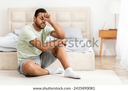 Upset young black man in pajamas sitting on floor by bed at home, thinking about something, looking for solution while 2024 crisis, leaning on his hand, panorama with copy space Royalty-Free Stock Photo #2435890081