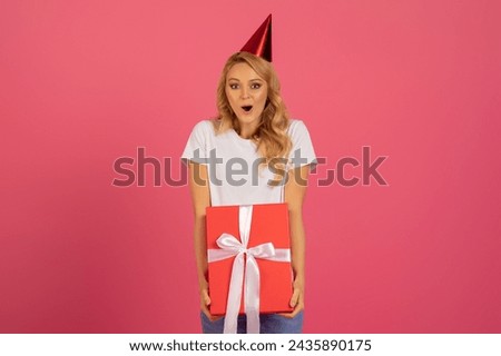 Happy Occasion. Studio portrait of surprised young European woman with blonde hair and festive hat, charming smile, holding wrapped present box, celebrating birthday on pink backdrop