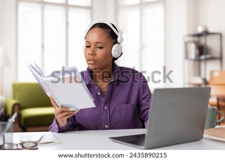 Concentrated black female student with headphones reviewing study materials and notes from textbook while simultaneously attending an online class on her laptop Royalty-Free Stock Photo #2435890215