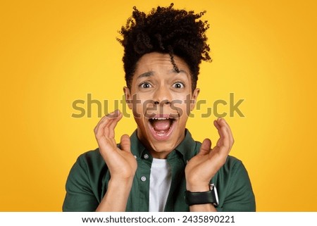 Closeup studio photo of emotional young african american man with fancy hairstyle screaming over yellow background, holding hands next to face, expressing amazement