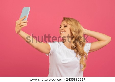 Young attractive blonde taking a selfie with her smartphone, posing for photo in casual white t-shirt, standing on pink studio backdrop. Concept of social media and online presence
