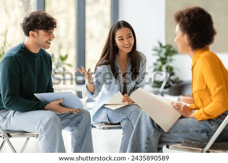 Three engaged diverse students sit in well-lit modern classroom, with one gesturing as they enthusiastically discuss the contents of their notebooks Royalty-Free Stock Photo #2435890589
