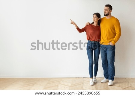 Look There. Happy caucasian couple pointing aside at copy space while standing against white wall background at home, young man and woman embracing and showing free place for your design, full length Royalty-Free Stock Photo #2435890655