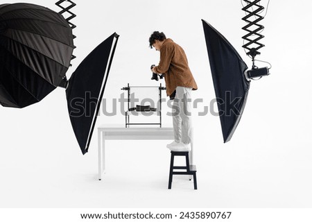 Focused male photographer stands on stool, meticulously adjusting his camera to capture the perfect studio shot, surrounded by professional lighting gear