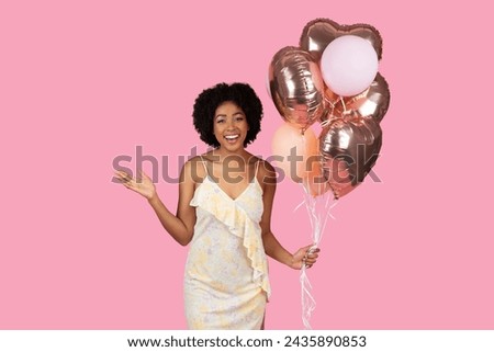 Vivacious cheerful millennial African American woman with curly hair joyfully presenting a bunch of rose gold and pink balloons, ready to celebrate on a pink background, studio Royalty-Free Stock Photo #2435890853