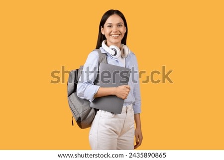 Content and ready-to-learn female student holds laptop while wearing headphones around her neck, showcasing preparedness and modern study habits against yellow background Royalty-Free Stock Photo #2435890865