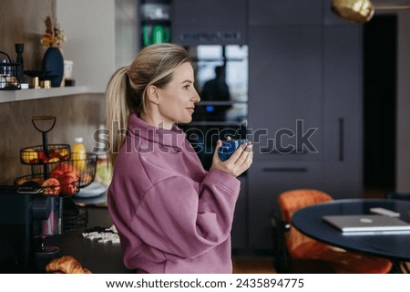 Woman enjoying cup of coffee at home after work, standing in kitchen wearing pink hoodie. Work-life balance for healthcare worker. Ear piercings.
