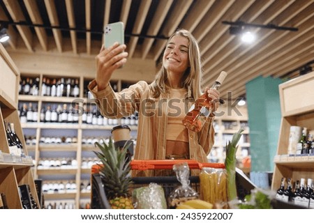 Young customer woman wears casual clothes hold wine bottle do selfie on mobile cell phone shopping at supermaket store shop with trolley cart buy product in hypermarket. Purchasing gastronomy concept