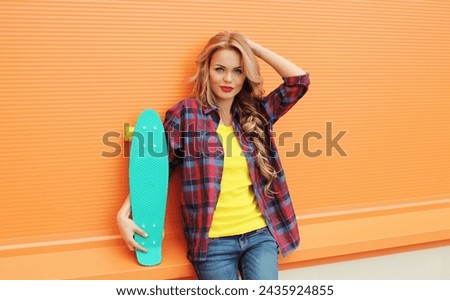 Portrait of stylish young blonde woman posing with skateboard on city street against colorful orange background