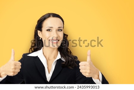 Businesswoman in black confident suit showing thumbs up gesture, yellow color background. Happy smiling gesturing brunette woman at studio. Business concept photo.