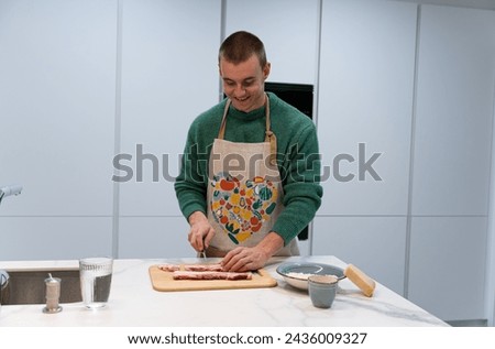 A man laughing is cutting meat on a cutting board in a kitchen. He is wearing a green apron with a heart design