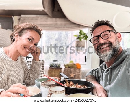 Happy couple eating together inside a camper van motorhome living the vanlife and travel adventure holiday vacation. Nomadic life. Cheerful man and woman taking selfie and smiling at the camera