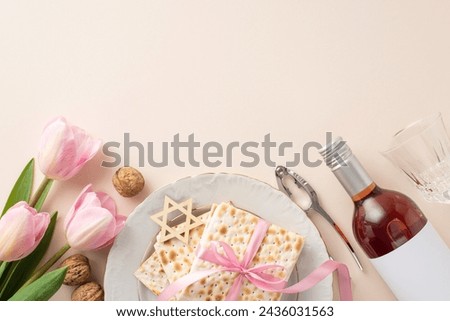 Top view picture of a Passover seder plate with matzah in a ribbon, a red wine bottle, a glass, nuts and nutcracker, star of David, and fresh tulips, on a pastel beige surface