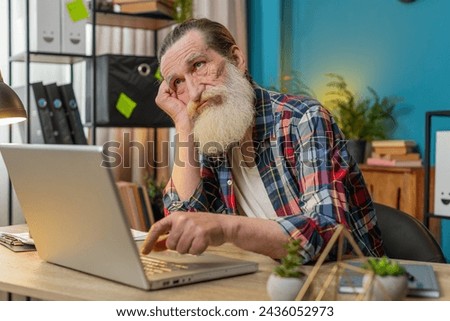 Overtired Caucasian senior man bored working on laptop while sitting in home office. Sleepy grandpa cannot concentrate on work, types lazily. Male freelancer is not interested in doing a project Royalty-Free Stock Photo #2436052973