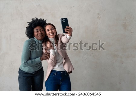 Two multicultural women making silly faces while taking photos with mobile phone.
