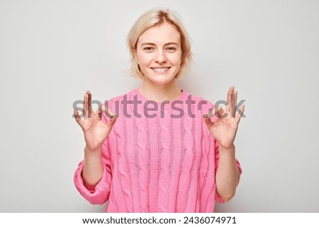 Smiling woman in pink sweater making OK sign with hand on a light background.