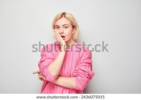 Surprised young woman in pink blouse with hand on cheek, white background.