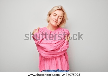 Portrait of a content woman in a pink sweater hugging herself against a gray background. Royalty-Free Stock Photo #2436075095