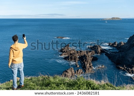 Man with his back turned in yellow sweatshirt and jeans on the cliffs of the Vadca coast taking a picture of the natural landscape on a sunny day with a clear blue sky.
