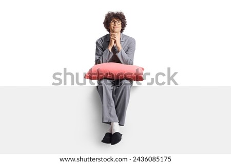 Young man in pajamas sitting on a blank panel and holding a pillow isolated on white background