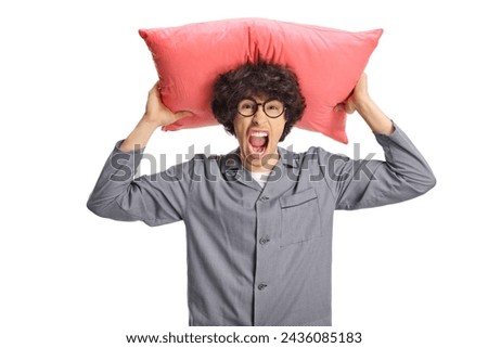 Angry young man in pajamas holding a pillow over head and screaming isolated on white background