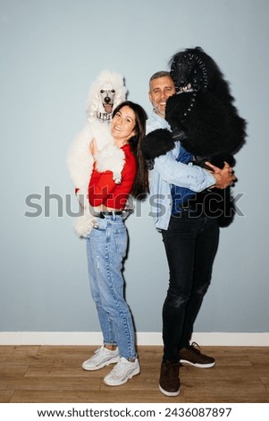 A man and a woman are posing for a picture with two poodle dogs. They are smiling and standing close together as they hold the dogs looking at camera