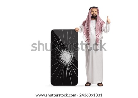 Saudi arab man in ethnic clothes leaning on a mobile phone with cracked screen and gesturing thumbs up isolated on white background