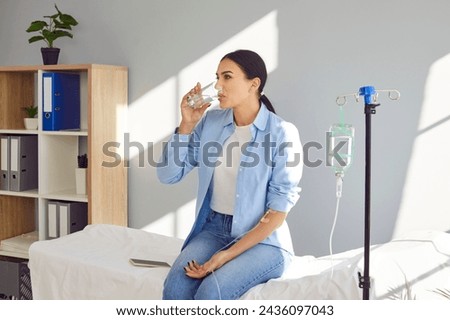 Young woman with sterile needle in arm sitting on medical bed by pole with IV drip bottle, drinking glass of water and receiving vitamin therapy infusion to boost immune system and suppress infection Royalty-Free Stock Photo #2436097043