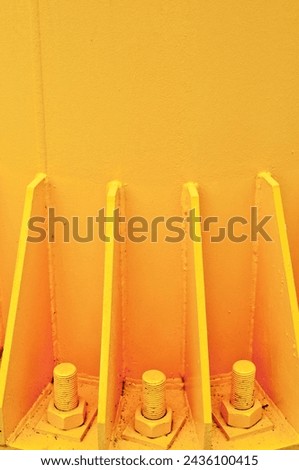 Close-up view of three large bolts anchored into yellow concrete surface, with metal supports painted in same color. Monochromatic setting, plain background.