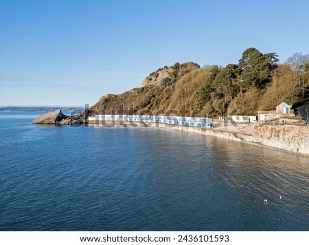 Aerial view of a long row of blue beach huts with trees and rocks behind and a calm blue sea at high tide at Meadfoot Beach in Torquay Devon.
