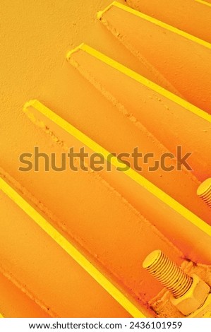 Close-up view of three large bolts anchored into yellow concrete surface, with metal supports painted in same color. Monochromatic setting, plain background.