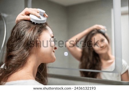 Young woman doing self hair scalp massage with scalp massager or hair brush for hair growth stimulating at home bathroom. Reflected view of the mirror. High quality photo