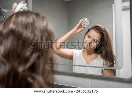 Young woman with dark hair doing self hair scalp massage with scalp massager or hair brush for hair growth stimulating at home bathroom. Reflected view of the mirror. High quality photo
