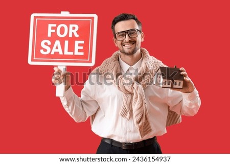 Portrait of male real estate agent with house model and FOR SALE sign on red background