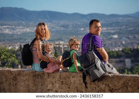 Family of four - mother, father, boy girl sit on viewpoint wall up high and turn to camera happy to visit Alhambra ancient castle on their summer journey to Granada, Spain Royalty-Free Stock Photo #2436158317