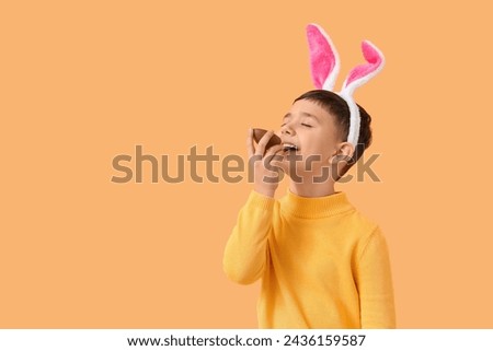 Cute little boy in bunny ears headband eating chocolate egg for Easter on orange background Royalty-Free Stock Photo #2436159587