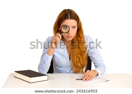 Portrait serious business woman teacher with glasses skeptically looking at you through magnifying glass sitting at desk isolated white background. Human face expression, body language, attitude