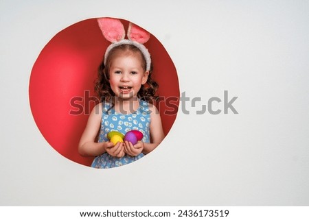 Cute little child wearing bunny ears on Easter day. Girl with painted eggs on white and pink background. 