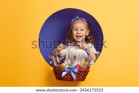 Cute little child wearing bunny ears on Easter day. Girl with painted eggs on bright purple and yellow background.  Royalty-Free Stock Photo #2436173523