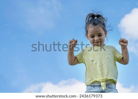 A little girl laughs with her arms raised under the bright summer sunher infectious laughter filling the air as the blue sky creates a perfect backdrop for this scene of pure joy and childlike happine Royalty-Free Stock Photo #2436179373