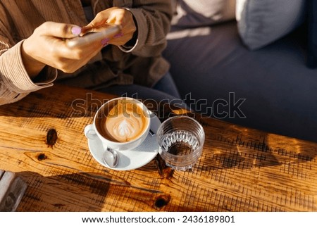 A girl holding phone and taking picture of cappuccino on the table coffee on the table 