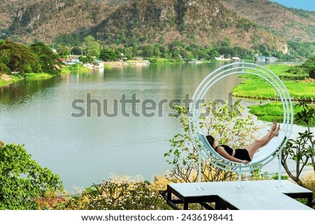Happiness woman relaxing on comfortable seat with beautiful view of nature at balcony on hill near riverbank. Female resting on curved seats on hillside terrace with mountains and river in background.