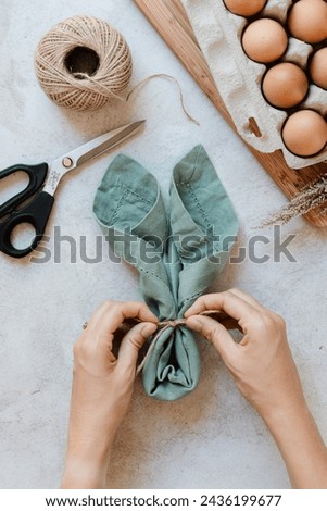 How to DIY a napkin folded in shape of bunny ears and an egg in the middle for an Easter themed celebration table setting, tying the string Royalty-Free Stock Photo #2436199677