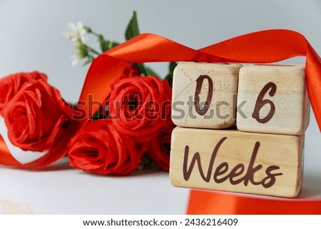 6 week gestational age milestone written on a wooden cube with red roses and ribbons, and a white background
