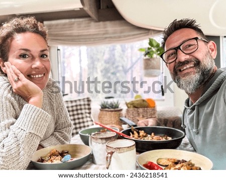 Happy couple eating together inside a camper van motorhome living the vanlife and travel adventure holiday vacation. Nomadic life. Cheerful man and woman taking selfie and smiling at the camera