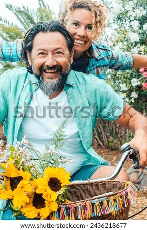 Young mature couple youthfully enjoy outdoor leisure activity together having fun and laughing. Man and woman riding a bike outside with flowers in background. Spring healthy sustainability lifestyle Royalty-Free Stock Photo #2436218677